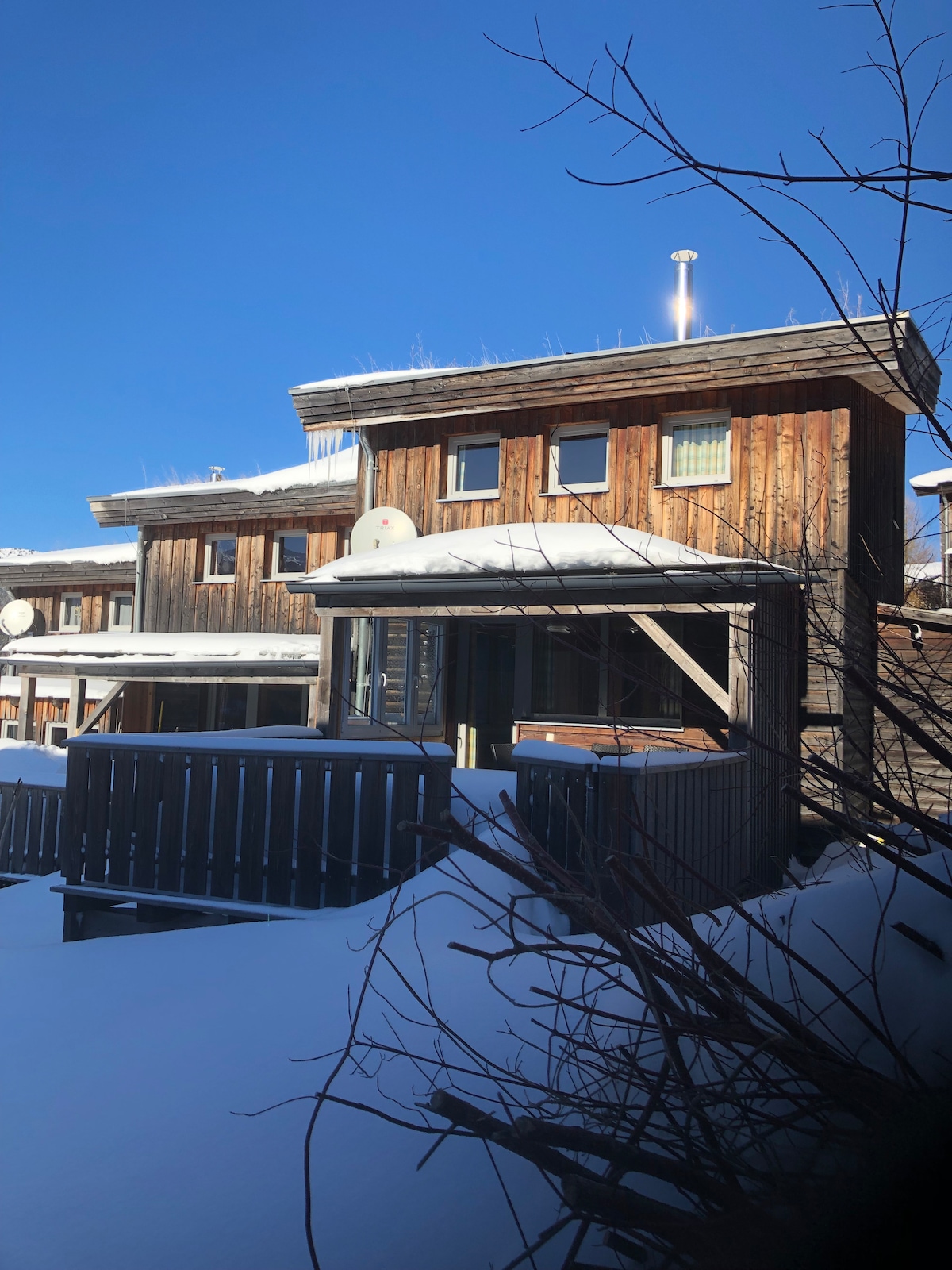 A wooden chalet is set against a clear blue sky, showcasing a blend of natural materials. Snow blankets the ground, with the structure featuring a covered terrace and a few windows, providing warmth to the winter landscape.