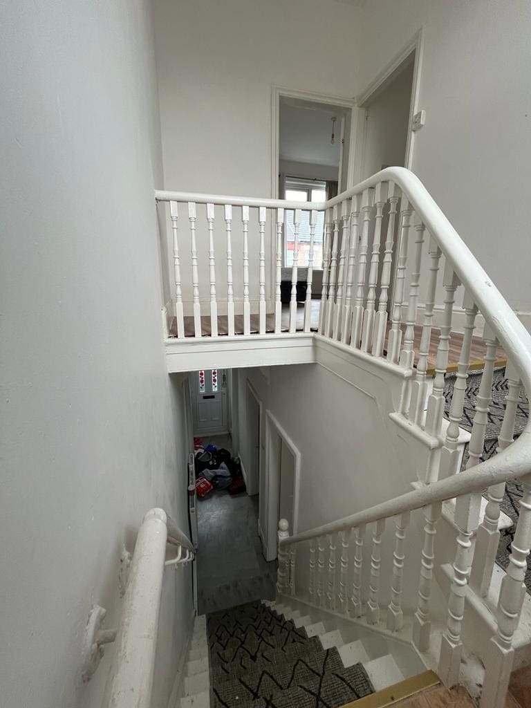 A bright staircase is visible, featuring white railings and a light-colored wall. The upper landing opens to a door, while the lower section leads to another entryway. A textured rug is placed at the bottom of the stairs, providing a cozy touch to the space.