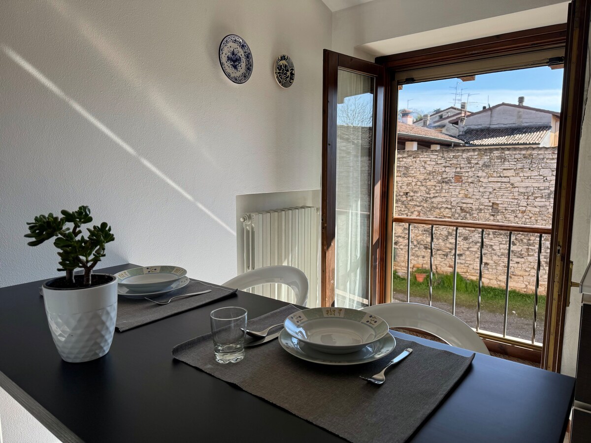 A dining area is presented with a dark table set for two, featuring plates and glassware atop grey placemats. Natural light enters through a window that overlooks a stone wall and the greenery outside, creating an inviting space.