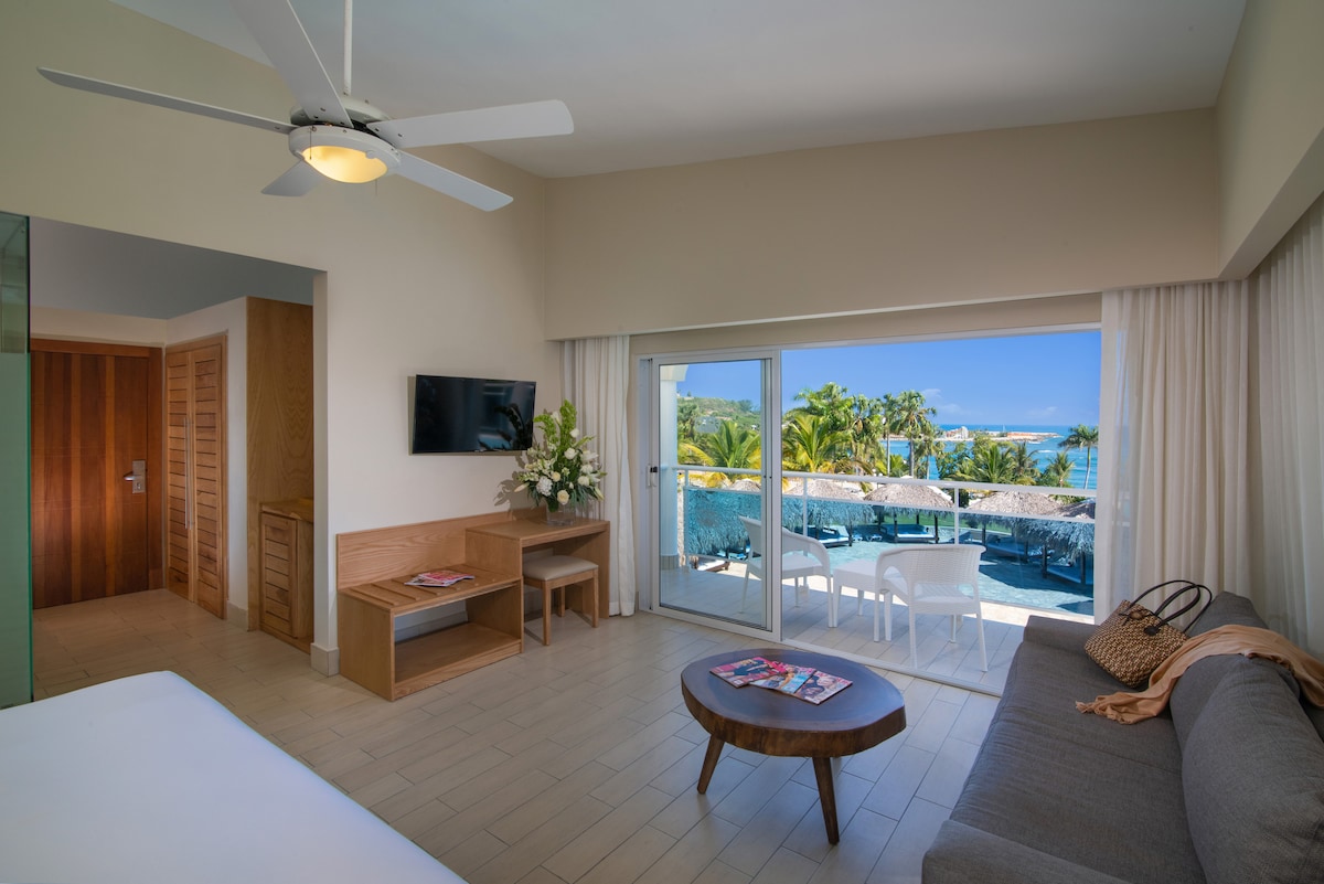 A bright and airy room is featured with a spacious bed, a cozy seating area, and a flat-screen TV mounted on the wall. Large glass doors lead to a balcony overlooking a tropical landscape, with outdoor seating providing a relaxing space to enjoy the view.