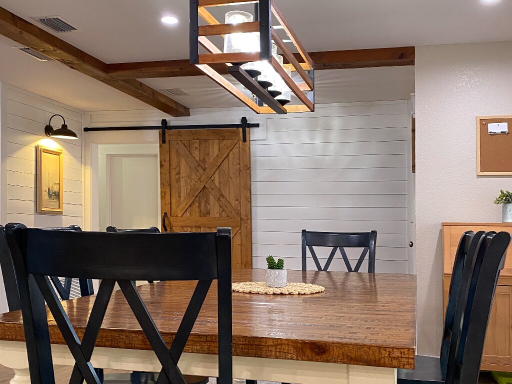 A dining area features a rustic wooden table surrounded by black x-back chairs. A linear chandelier hangs above, illuminating the space. A sliding barn door and light-colored walls add to the farmhouse aesthetic.