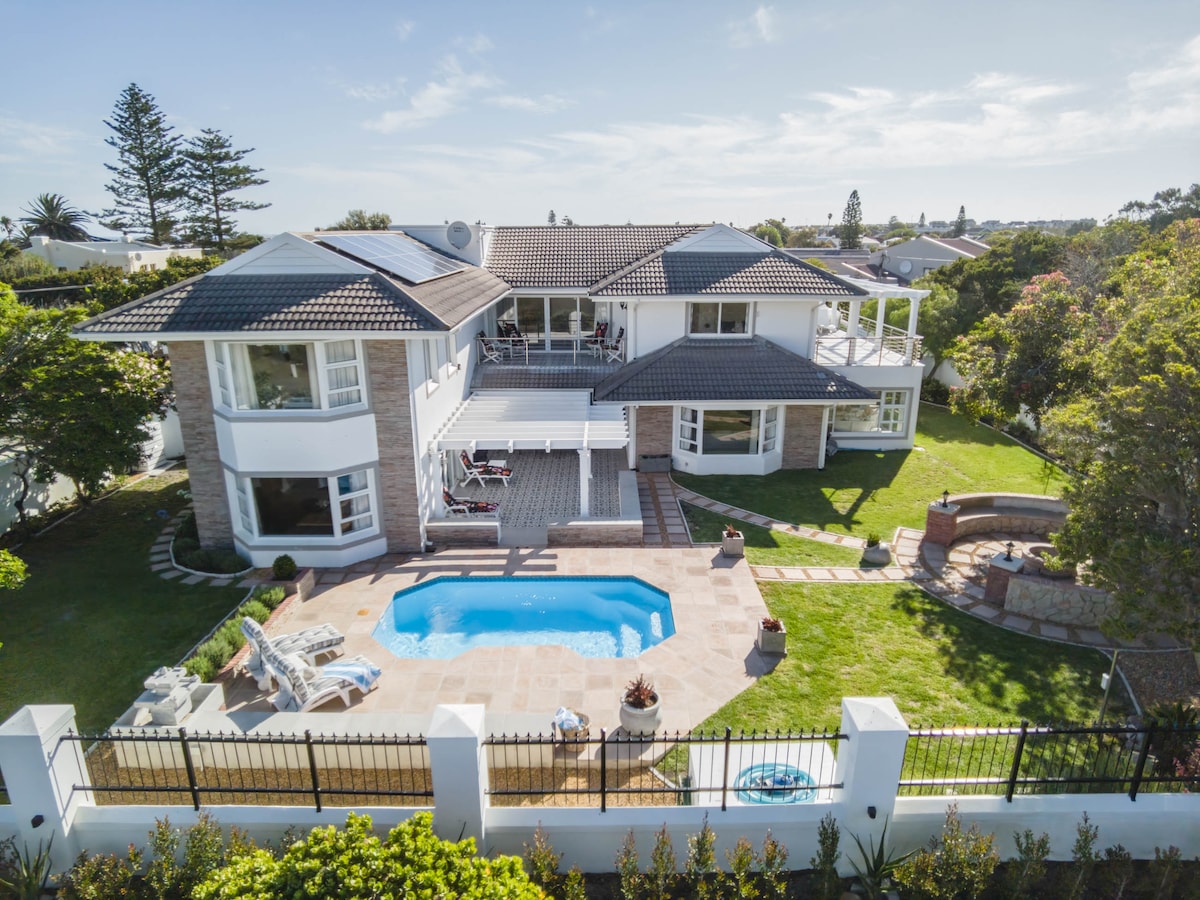The expansive property features a well-maintained lawn with a pool bordered by a patio, offering outdoor furniture for relaxation. A fire pit can be seen in one corner, with the two-story house displaying multiple balconies and large windows that allow natural light to flood the interior.