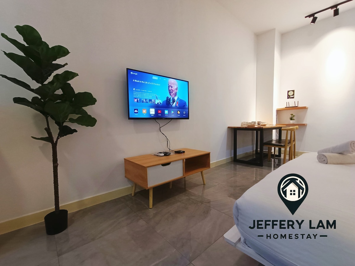 A well-lit studio space is shown, featuring a mounted flat-screen television against a white wall. A wooden TV stand complements the modern decor. A small dining table is positioned in the background along with a potted plant, contributing to the open, uncluttered environment.