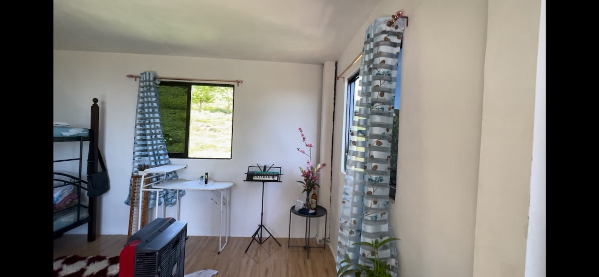 The room features natural light from large windows framed by patterned curtains. A compact table stands beside a chair, while a flower vase adds a touch of nature. A cozy sleeping area is visible in the corner, complemented by a cool-toned rug.