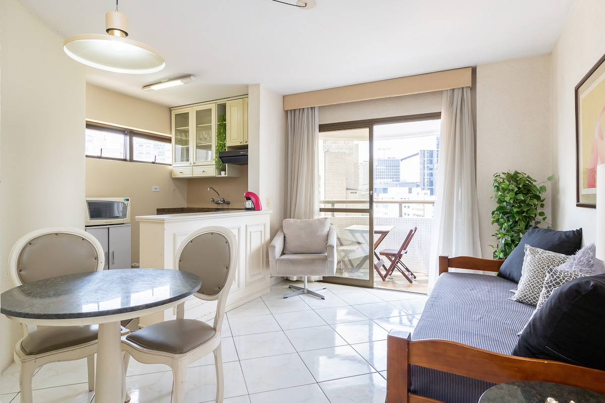 A modern living space is presented with a compact kitchen, featuring light green cabinets and essential appliances. A cozy seating area is shown, complemented by a small dining table. A balcony is visible through large glass doors, offering a glimpse of the outdoor view.