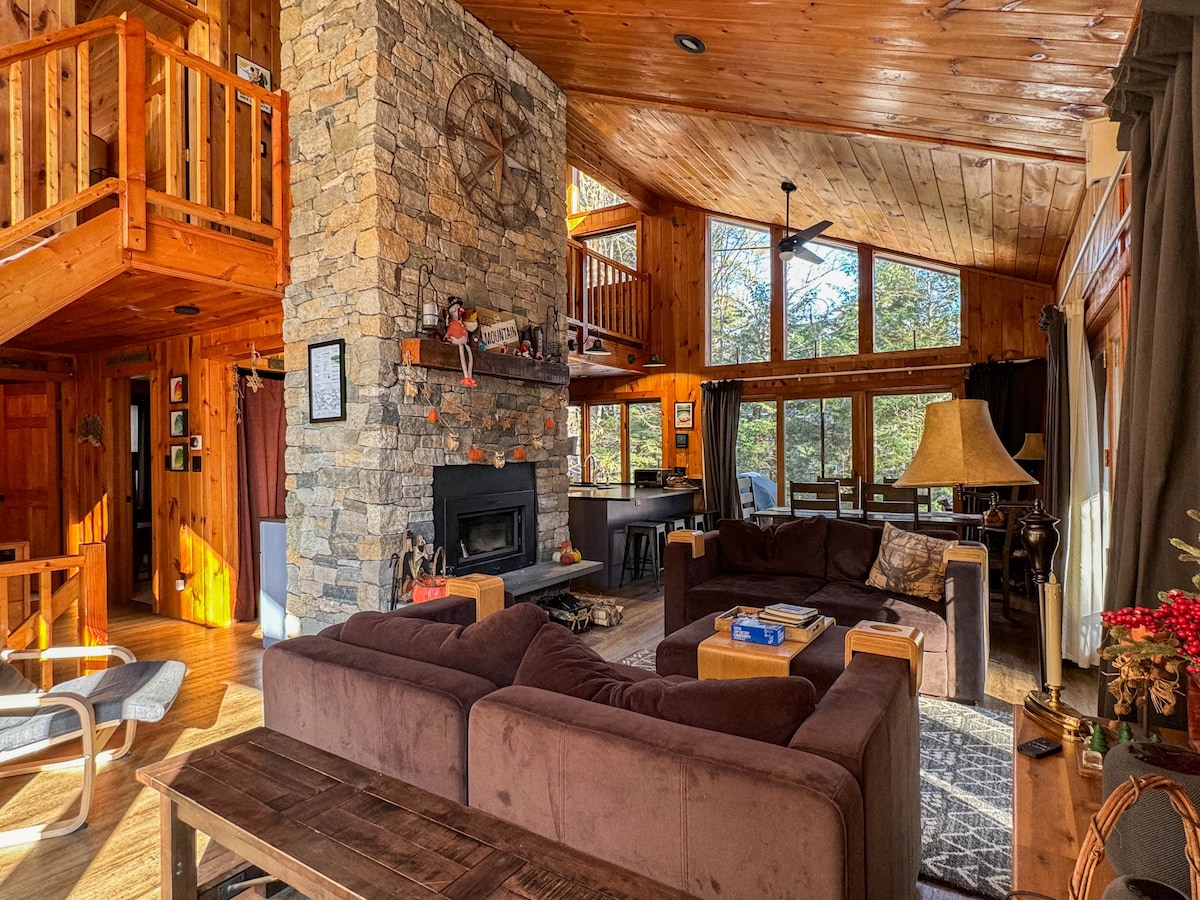 A spacious living area features a stone fireplace as the focal point, surrounded by large windows that invite natural light. Comfortable seating is arranged around a central coffee table, with a cozy rug underfoot. Wooden beams and warm tones reflect the rustic ambiance of the space.