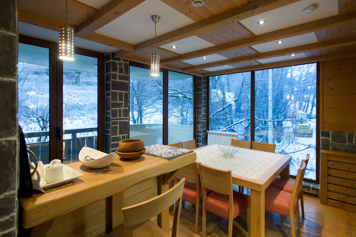 A spacious dining area is illuminated by recessed lighting, featuring a large wooden table surrounded by chairs. Floor-to-ceiling windows showcase a snowy landscape outside, and a counter with decorative items adds to the inviting atmosphere.