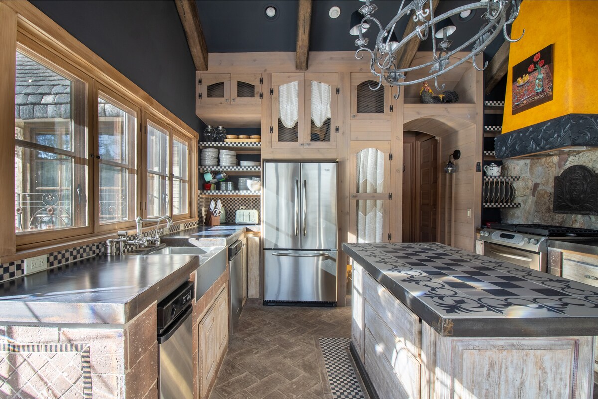 The kitchen features an open layout with wooden cabinets and a decorative island. Full-height windows provide natural light, while stainless steel appliances create a modern touch. Black and white patterned tiles accentuate the countertop, complemented by a rustic charm of wood accents throughout the space.