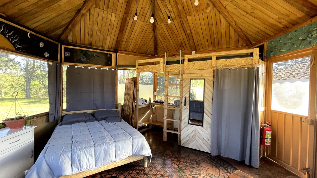 A tranquil interior space features a spacious bed with a soft blue blanket at its center. Wooden walls and a high, peaked ceiling create an open atmosphere. Natural light enters through large windows, while grey curtains add a touch of privacy.