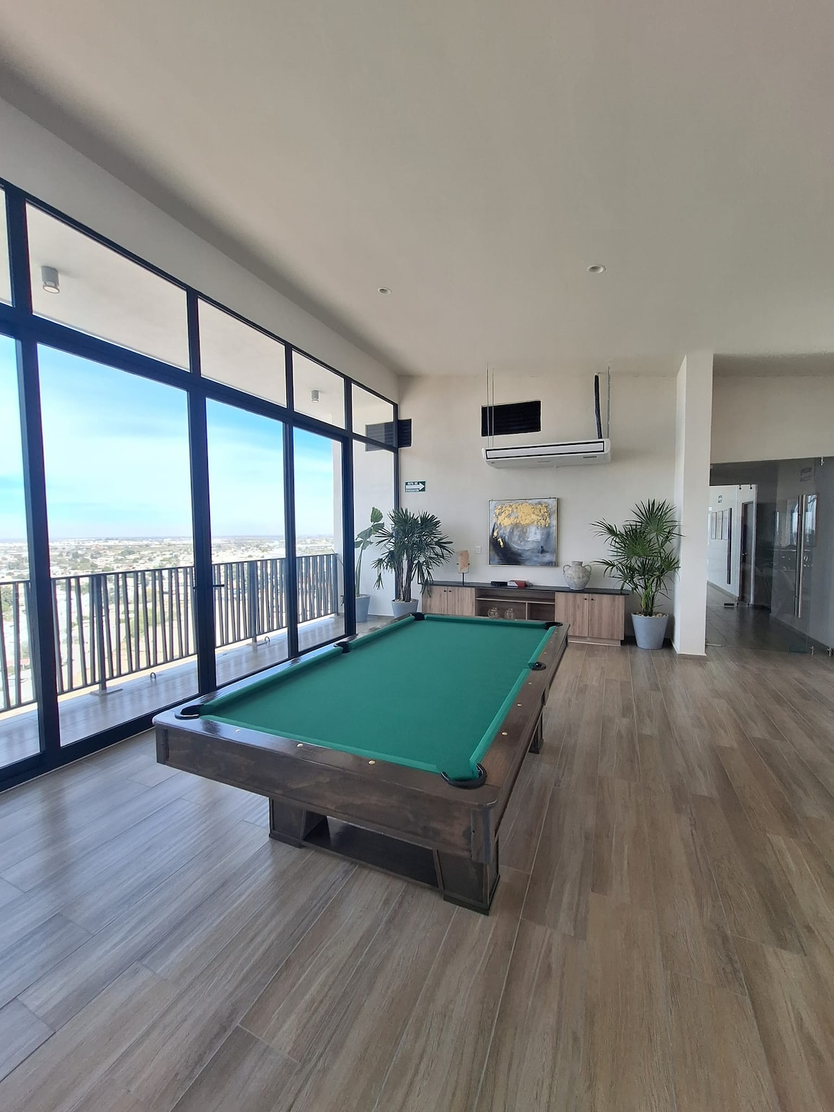 A spacious game room features a billiards table with a green felt surface. Large floor-to-ceiling windows provide panoramic city views and ample natural light. Potted plants add a touch of greenery, complementing the light wood flooring and modern design elements.