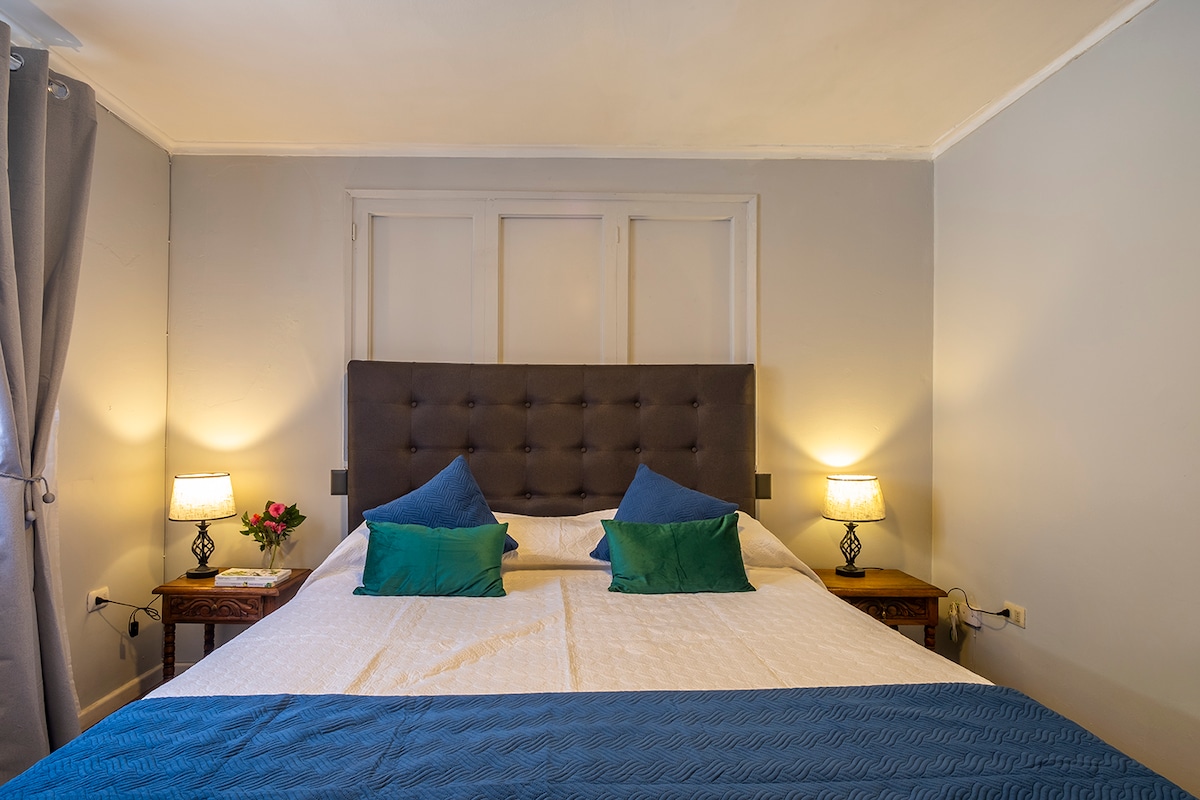 A spacious bed is adorned with light-colored linens and topped with teal and blue accent pillows. Warm light is provided by two bedside lamps on wooden tables. Two curtains frame a window, complementing the neutral-toned walls and adding a calming effect to the room.