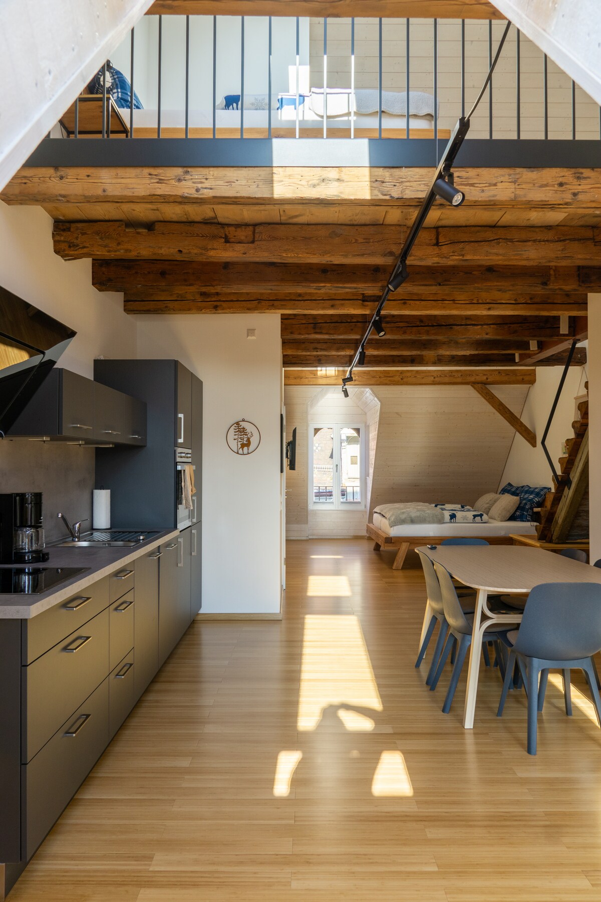 An open layout studio features a fully equipped kitchen with modern appliances on one side, while natural light floods the space through large windows. A dining table for six is positioned nearby, and a staircase leads to the upper area where an additional king-size bed awaits.