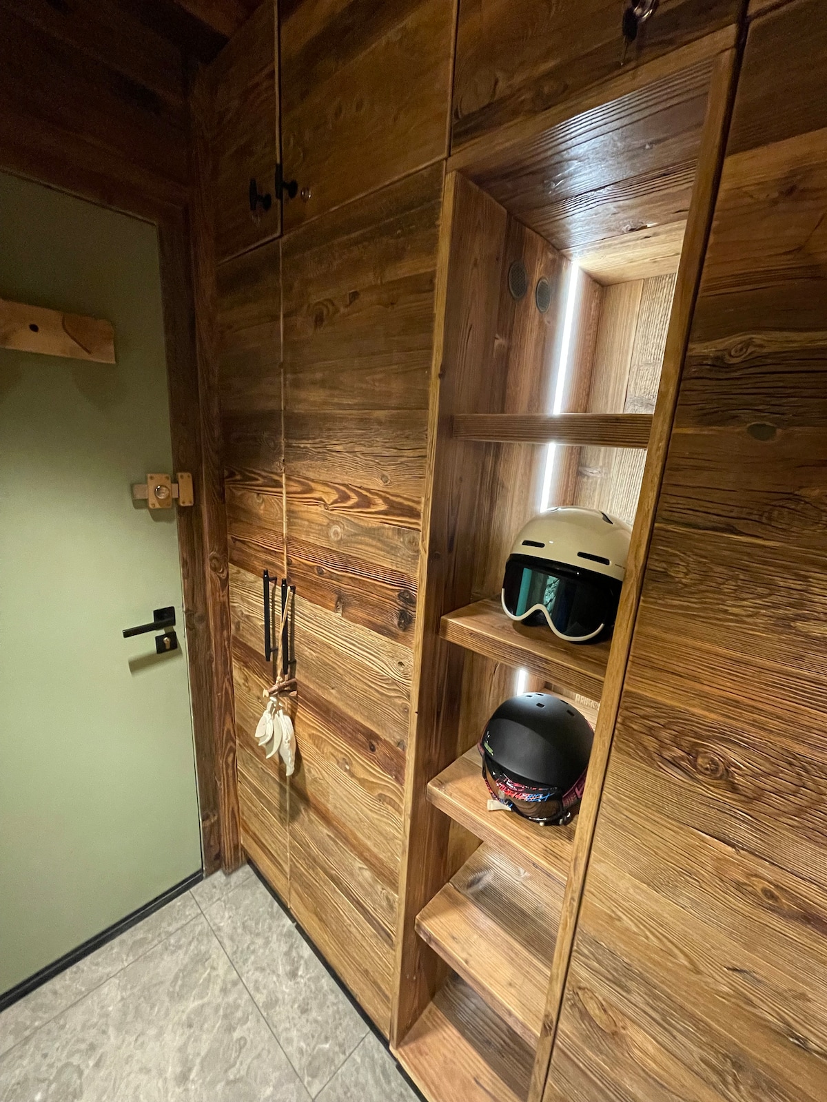 A well-organized entryway features wooden cabinetry with ample storage and open shelving. Ski helmets are displayed on the shelves, while a door with a simple handle provides access to the apartment. The flooring is covered with sleek grey tiles, enhancing the overall modern design.
