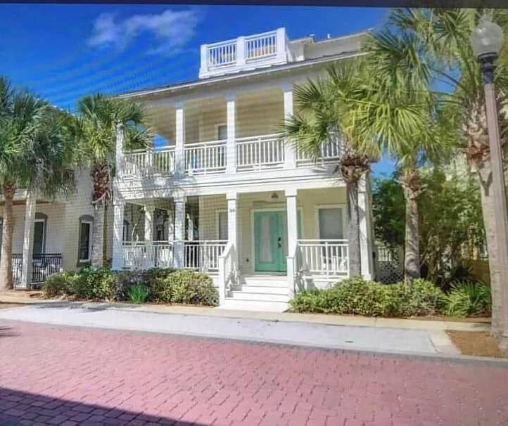 A charming two-story house features a light-colored façade with a teal front door and ample balcony space. Lush palm trees frame the entrance, while brick paving leads to the property. The home is surrounded by greenery, enhancing its inviting exterior.