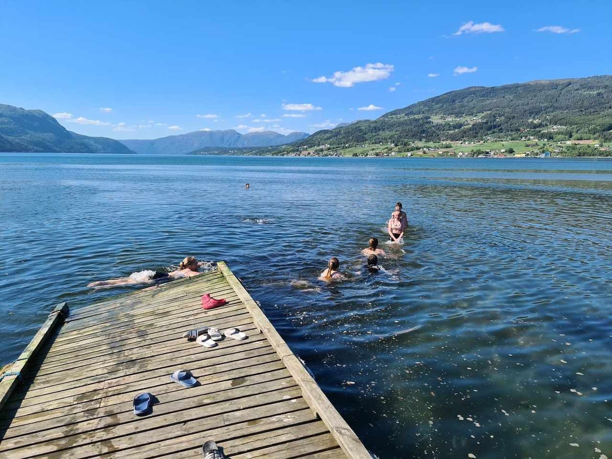 A wooden pier extends into a calm body of water, where several individuals can be seen swimming. The surrounding landscape features rolling hills and mountains under a bright blue sky, creating a serene natural backdrop.