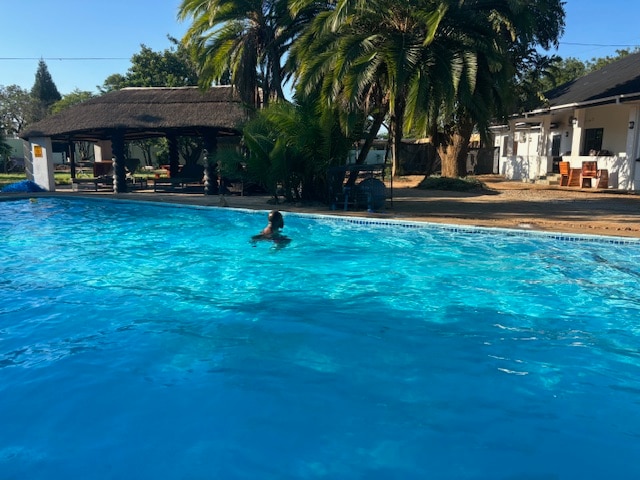 A serene swimming pool is visible, with a person swimming in its clear water. Surrounding palm trees and a thatched-roof gazebo provide shade. In the background, a well-maintained garden area features seating arrangements, creating a relaxing environment.