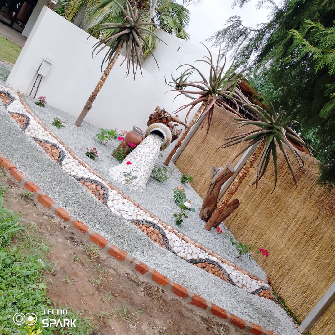 A landscaped garden is featured with decorative gravel paths, vibrant plants, and palm trees. A stone water feature is positioned prominently, creating a serene focal point. The area is enclosed by bamboo fencing, contributing to a sense of privacy and calm.