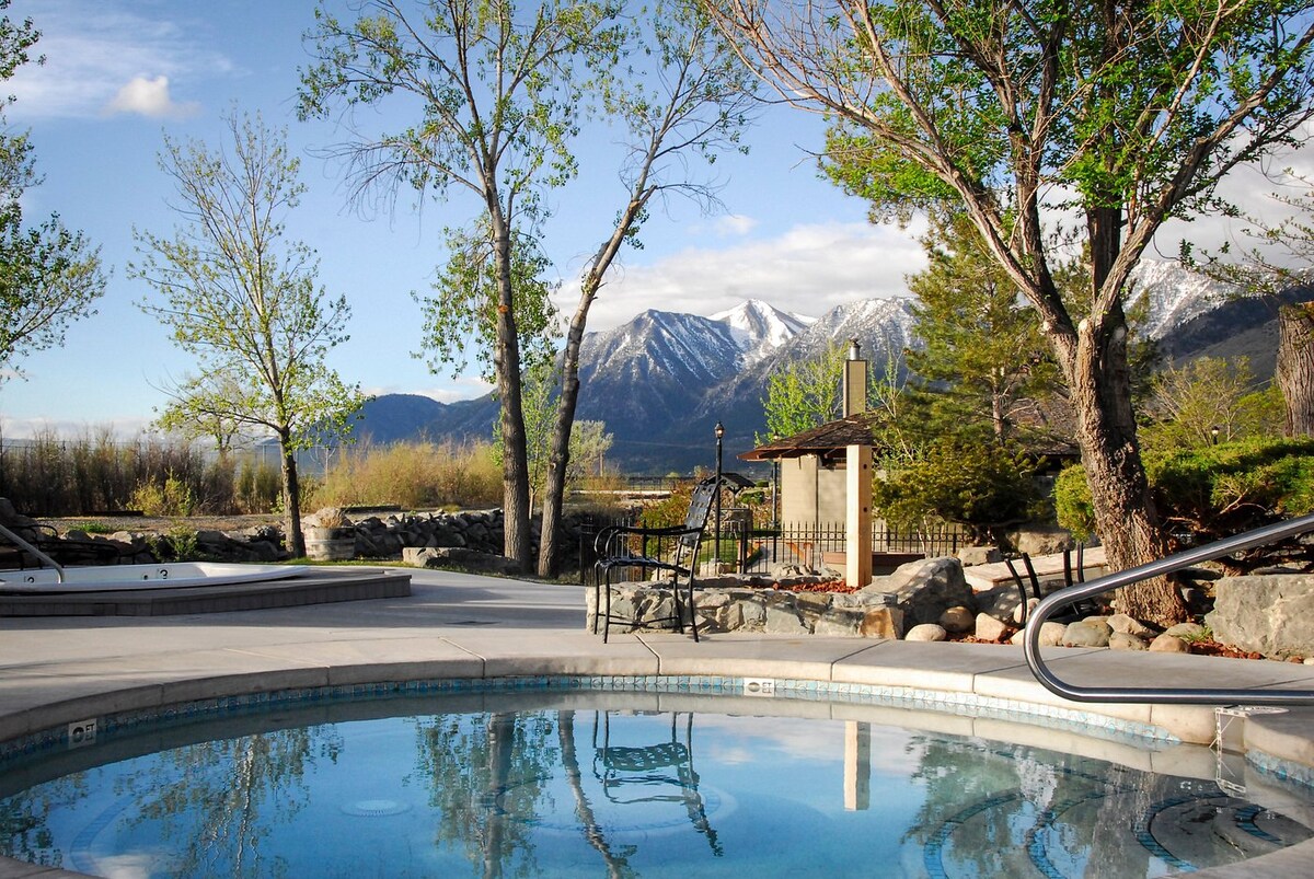 A sparkling hot tub surrounded by a concrete patio features views of the mountains in the background. Lush trees line the area and a barbecue station is visible nearby, creating a relaxing outdoor space for guests.