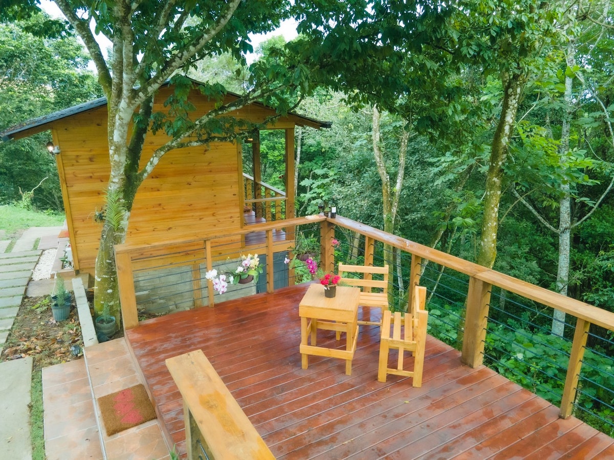 A wooden deck is surrounded by lush greenery, featuring a small table accompanied by two chairs. The rustic cabin is visible behind, complemented by natural wooden textures and an inviting outdoor space.