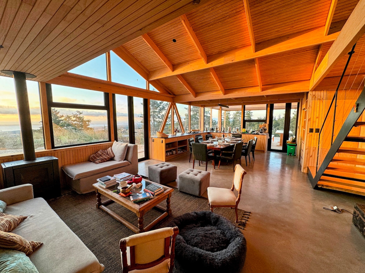 An open living space features large windows allowing for natural light and scenic views of the outdoors. A comfortable seating area with a mix of furniture is complemented by a dining table in the background, framed by warm wood finishes and a staircase.