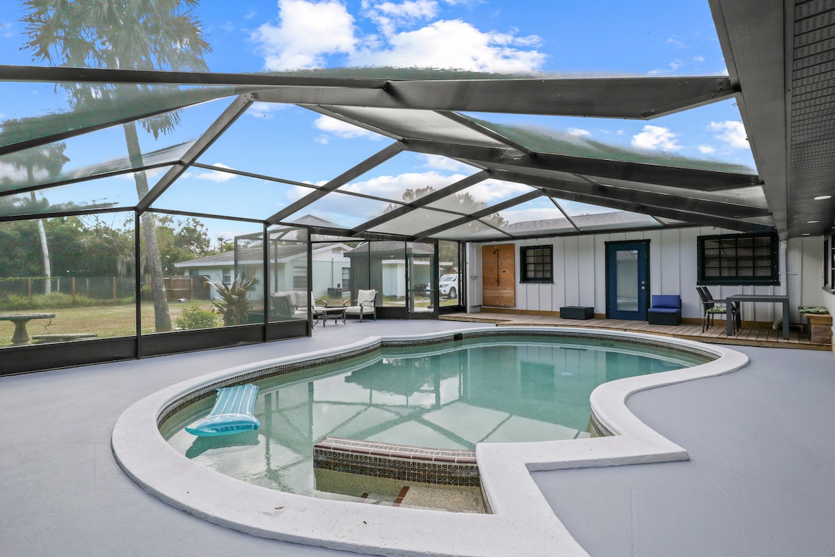 An outdoor pool area is enclosed with a screened lanai, maximizing comfort. The pool features a shallow ledge and a spa section, surrounded by modern seating arrangements. Clear skies and palm trees add to the serene ambiance of the space, inviting relaxation.
