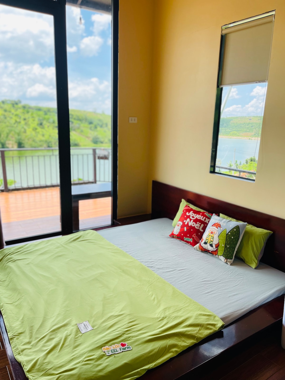 A spacious sleeping area features a bed with a light green blanket layered over a neutral-colored base. Pillows with festive designs are placed at the head of the bed. Large glass doors provide a view of the surrounding landscape and access to a balcony.