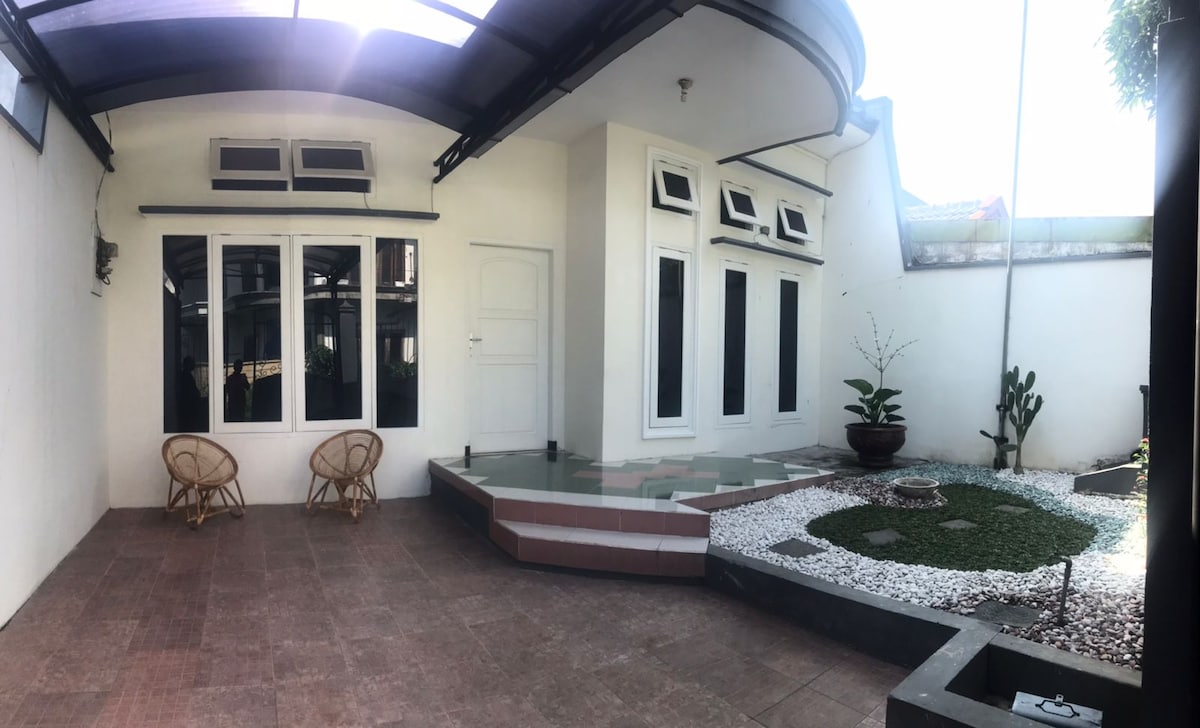 An outdoor area features a small gravel garden with decorative stones, surrounded by greenery. Two rattan chairs are positioned on the tiled patio, facing a white wall with large windows that allow natural light to fill the space.