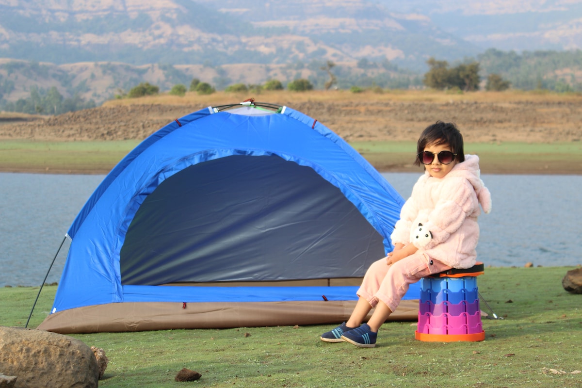 A bright blue tent is pitched near a body of water, set against a backdrop of rolling hills. A child is seated on a colorful stool, wearing a soft pink sweater and light pants. The ground is a mixture of green grass and rocks.
