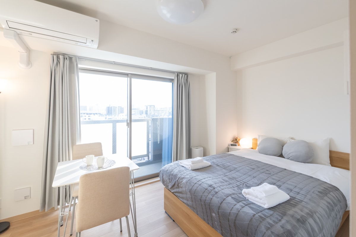 A well-lit room features a spacious bed with light bedding and soft pillows. A small dining table with chairs is positioned nearby. Large windows provide a view of the city, and light curtains create a bright atmosphere.