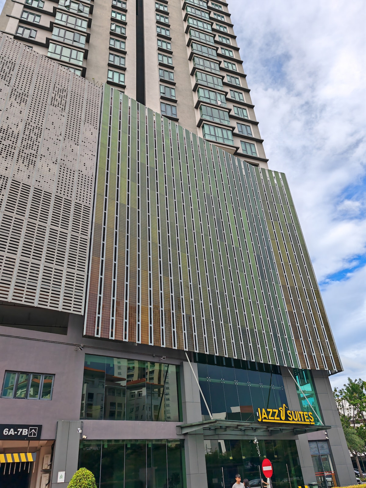 A modern building exterior is showcased, featuring a striking perforated façade with a variety of colors. The entrance to Jazz Suites is visible, marked by a welcoming sign, while a tall structure expands above, framed by a blue sky with scattered clouds.