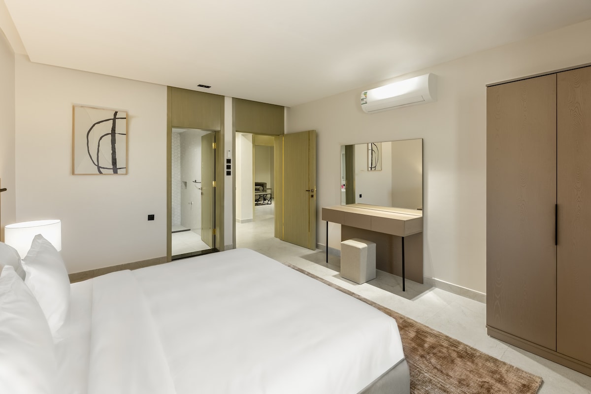 A spacious bedroom is furnished with a large bed, complemented by a contemporary vanity set and a modern wardrobe. Soft lighting emanates from a bedside lamp, while a neutral color palette creates a serene environment. An en-suite bathroom is partially visible through the open door.