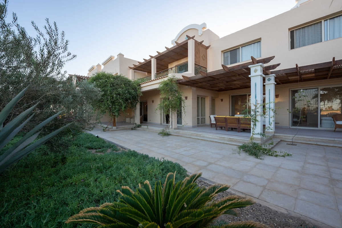The exterior of the townhouse features a spacious patio area with comfortable seating beneath a wooden awning. Lush greenery, including ornamental plants and trees, enhances the outdoor space, while the minimalist architecture offers a modern appeal. Natural light highlights the clean lines of the structure.