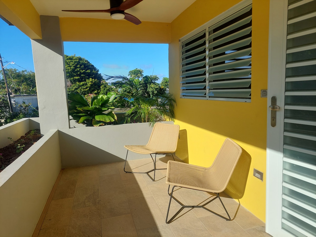 A bright outdoor space features two minimalist chairs positioned on a tiled floor. Large windows with adjustable slats allow natural light to fill the area, while vibrant yellow walls contrast with lush greenery visible in the background, creating a refreshing atmosphere.