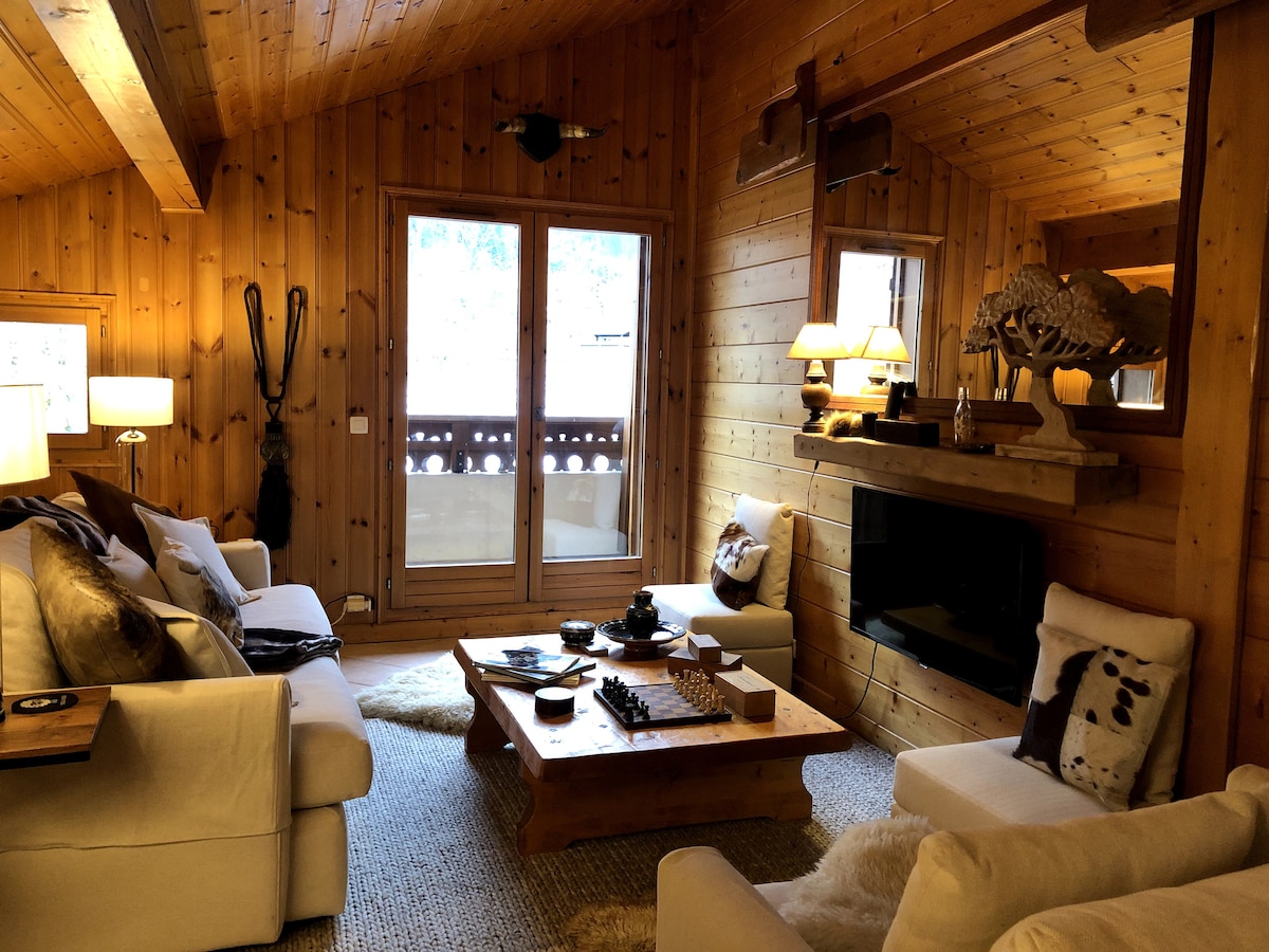 A warm living area is enhanced by wooden paneling and vaulted ceilings. Soft seating arrangements flank a central coffee table adorned with decorative items. Natural light enters through glass doors, which open to a balcony. A wall-mounted television is positioned above a console.