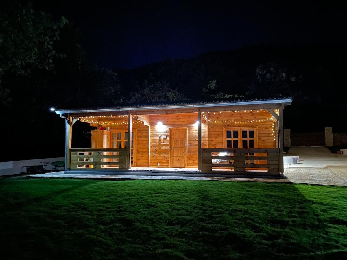 A warmly lit wooden lodge is showcased at night, featuring a charming exterior with string lighting along the porch. The structure features large windows, allowing glimpses of the inviting interior. Surrounding the lodge, a well-maintained lawn enhances the peaceful ambiance.
