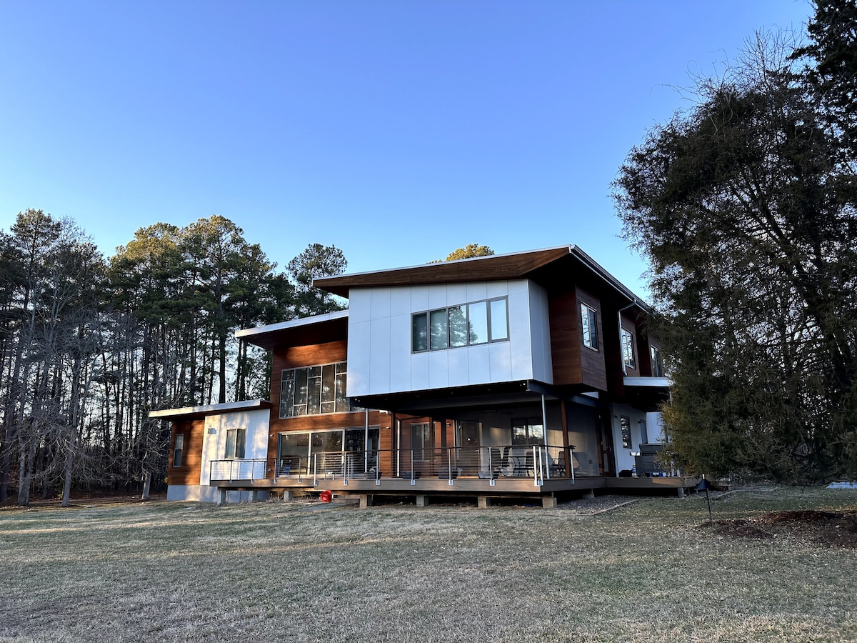 A modern home is set on a grassy lot, surrounded by tall trees. The structure features a mix of wood and white paneling, with a large deck accessible from multiple doors. Clear skies create a serene backdrop for this peaceful dwelling.