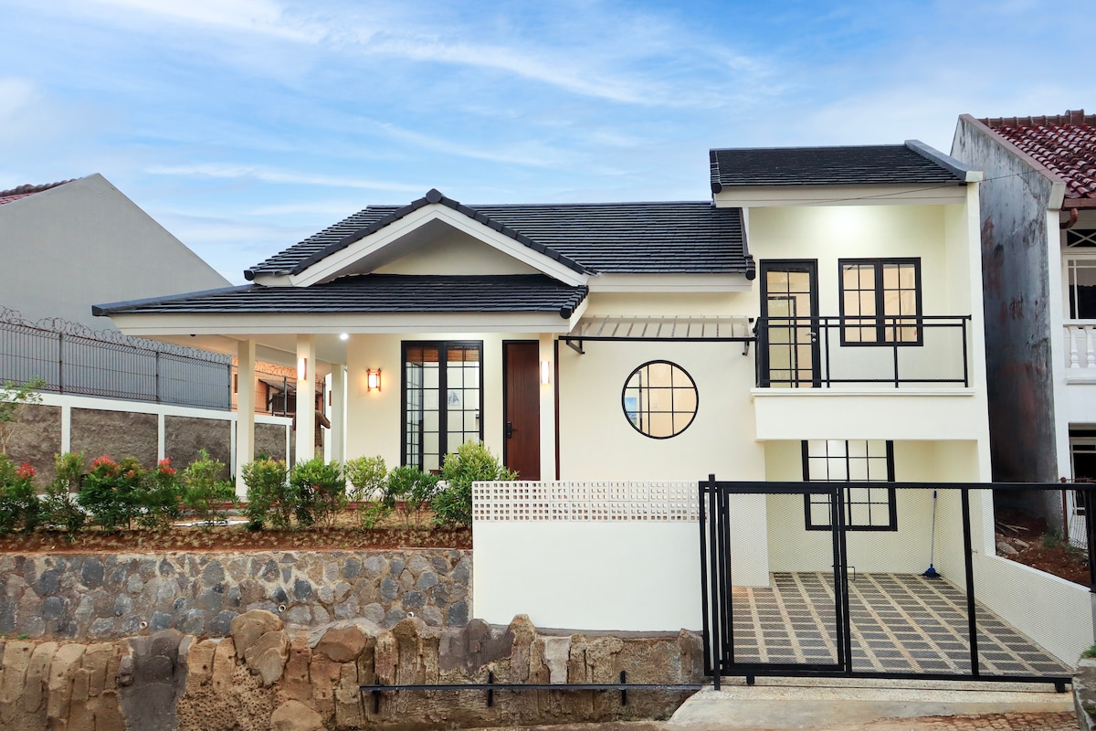 The villa features a modern architectural style, with a black roof and a light-colored exterior. Large circular windows provide natural light. A small garden with greenery is visible in front, alongside a paved area fenced for privacy.