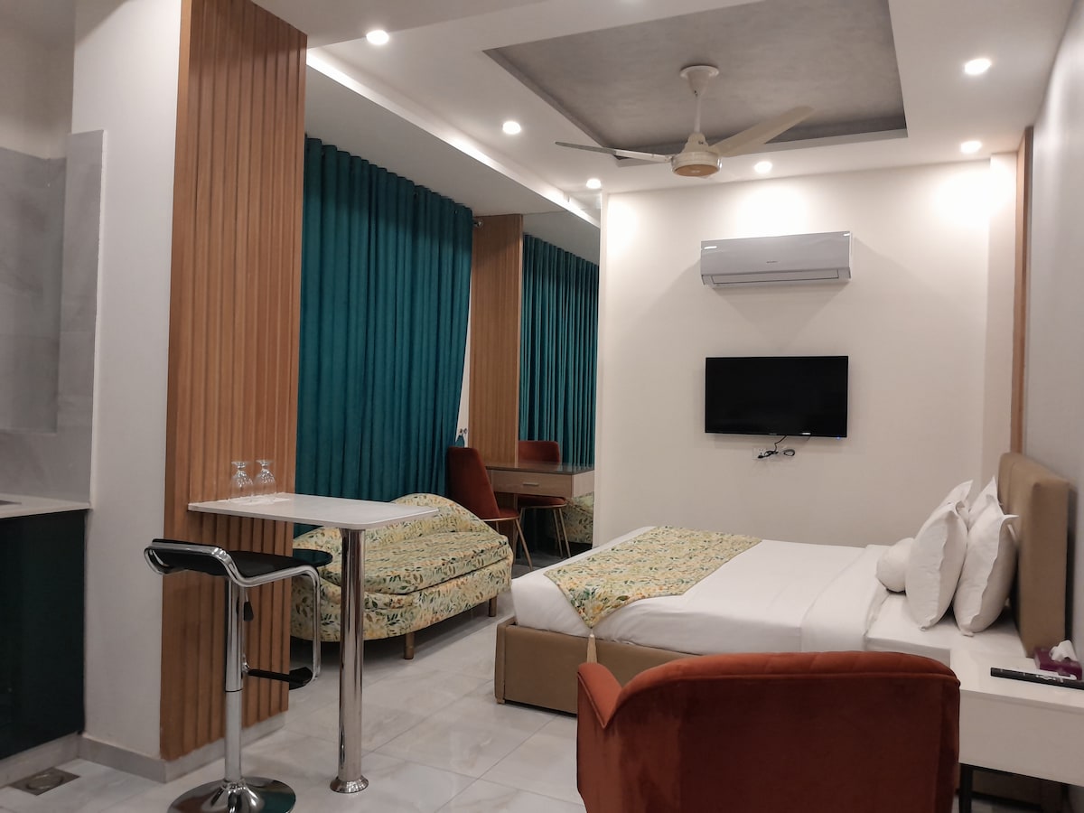 A modern studio space features a spacious bed complemented by a decorative throw. Two additional beds are positioned in the background. A small dining table and bar stools are visible, along with a wall-mounted television. Rich turquoise curtains frame the windows, enhancing the room's decor.