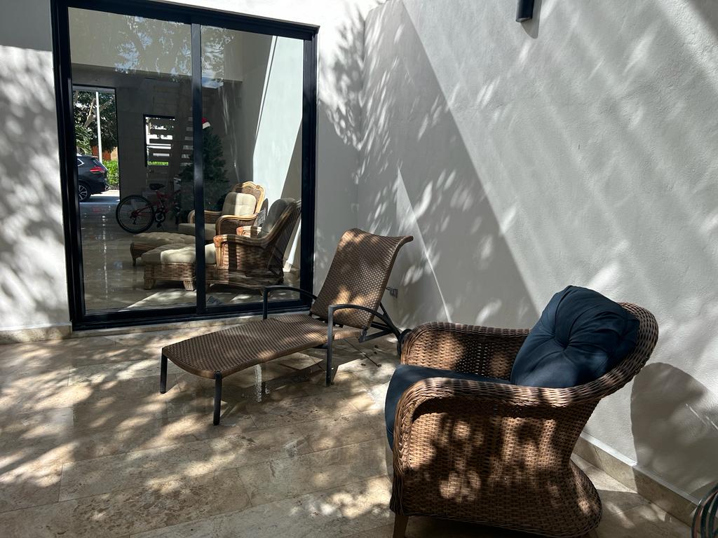 A shaded outdoor seating area features two comfortable wicker chairs and a lounge chair, situated on a polished stone surface. Light patterns created by nearby foliage add visual interest to the space, while large glass doors provide access to the interior.