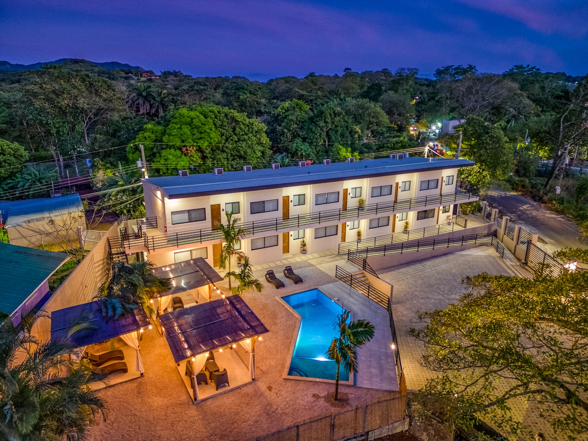An aerial view captures a modern condominium building surrounded by lush greenery. A private pool is seen, complemented by lounge chairs and shaded seating areas. The well-lit exterior highlights the inviting layout of the property, presenting an ideal setting for relaxation.