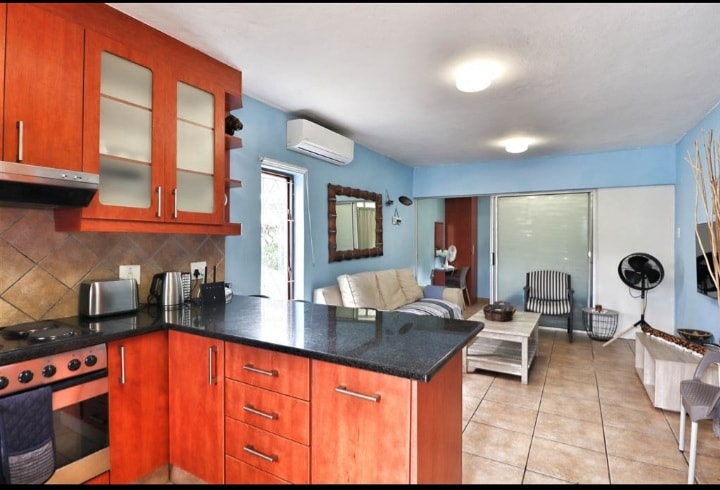 An open-plan kitchen and lounge area is depicted, featuring wooden cabinetry and modern appliances. A comfortable seating arrangement is visible in the lounge, complemented by a large mirror and soft blue walls. Natural light enters through a glass door leading to the outdoor space.