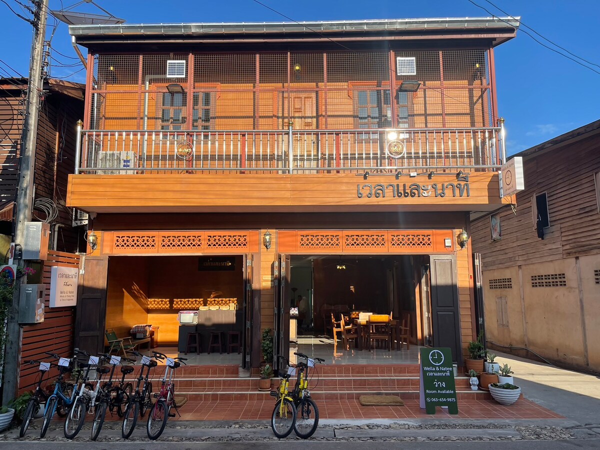 A traditional wooden exterior is showcased, featuring a two-story building with a welcoming balcony. The ground level is open, revealing a dining space with wooden furnishings. Bicycles are parked in front, hinting at leisurely exploration opportunities in the surrounding area.