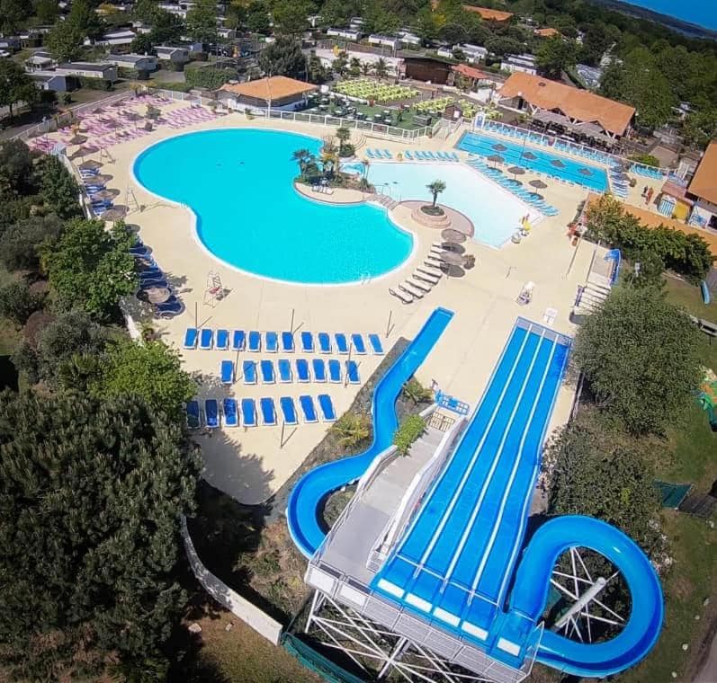 A spacious outdoor pool area features a large, irregularly shaped swimming pool surrounded by sun loungers and colorful umbrellas. A water slide is visible, leading into a shallow pool, with well-maintained gardens and buildings in the background.