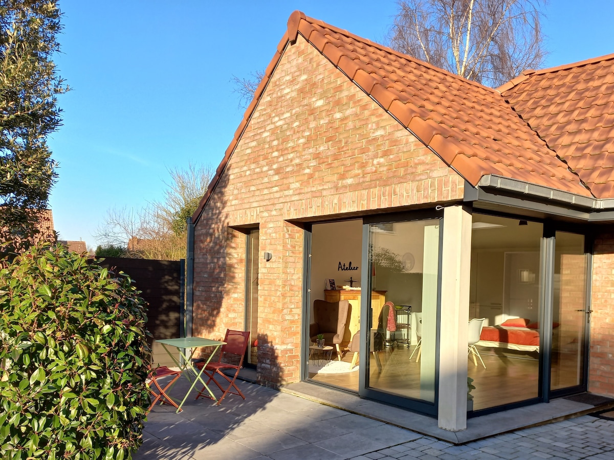 The exterior of a modern brick residence features a sloped roof and large glass doors that open to an inviting interior. A small table with two chairs is placed on the patio amidst greenery, creating a serene outdoor space.