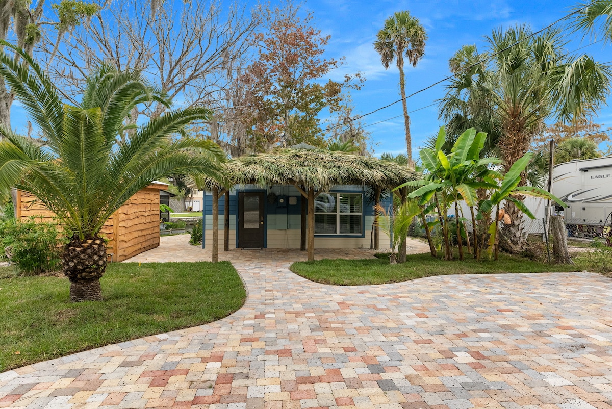 A modern shack is nestled among a lush landscape featuring palm and banana trees. The exterior showcases a thatched roof and large front windows. A paved pathway leads to the entrance, surrounded by well-maintained grass and tropical plants.