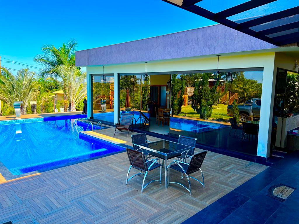 An outdoor area features a large, inviting swimming pool surrounded by sun loungers and a dining table with chairs. Expansive glass walls provide views of the lush greenery, enhancing the open-air feel. The patio is complemented by a shaded area for relaxation.