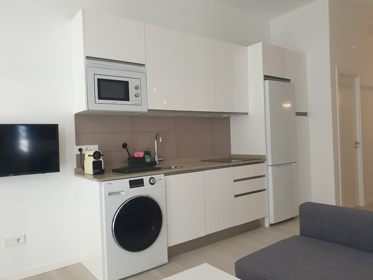 A modern kitchen area is equipped with sleek white cabinetry and a stainless steel refrigerator. Appliances include a microwave, a washing machine, and essential kitchenware displayed on the countertop. A wall-mounted television is visible, providing convenience for leisure activities.