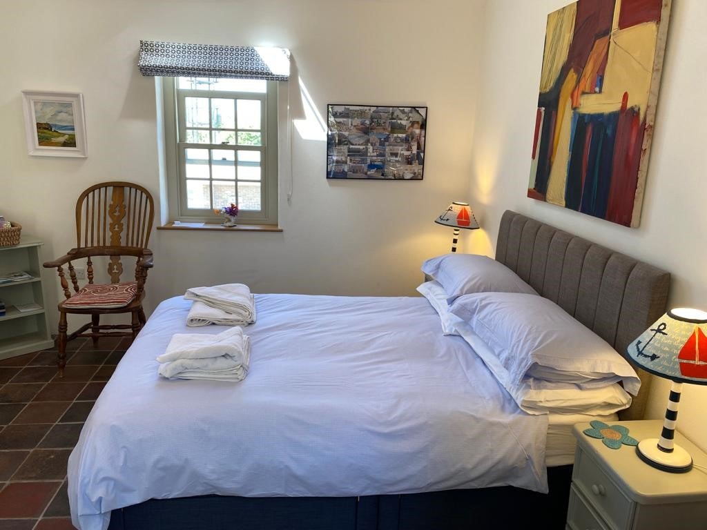 A cozy sleeping area is presented with a spacious bed dressed in crisp white linens. Flanking the bed, two bedside lamps provide soft illumination. A window allows natural light to enter, while a colorful wall art piece adds character to the room.