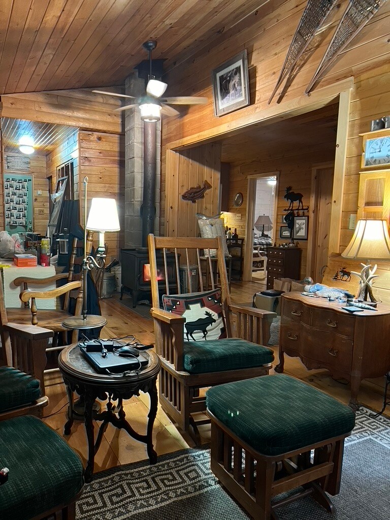 A spacious living area is highlighted by wooden walls and a warm atmosphere. Several wooden chairs surround a small table, while a standing lamp contributes soft lighting. A vintage fan is mounted on the ceiling, and rustic decor items are displayed throughout the space.