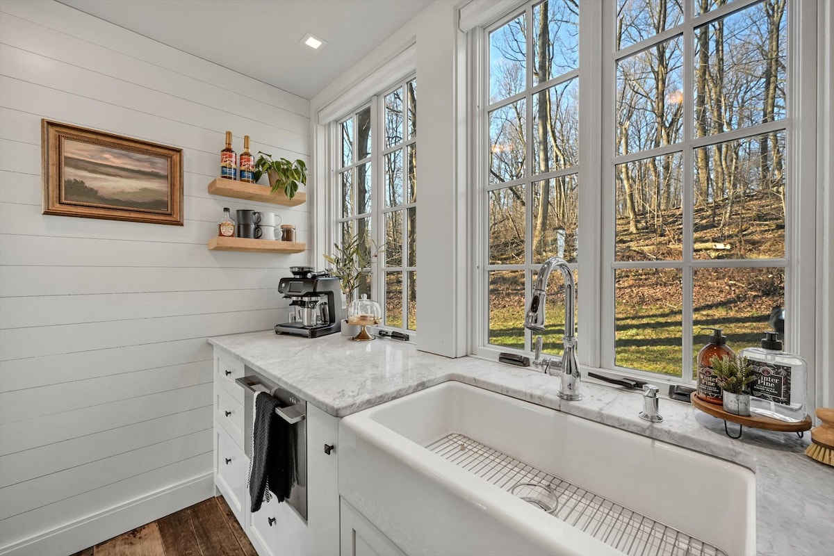 A bright kitchen is featured with large windows offering views of the serene wooded surroundings. Quartz counters and white cabinetry complement the space. A farmhouse sink is positioned beneath the window, and a coffee station is set up on the counter with a machine and supplies.